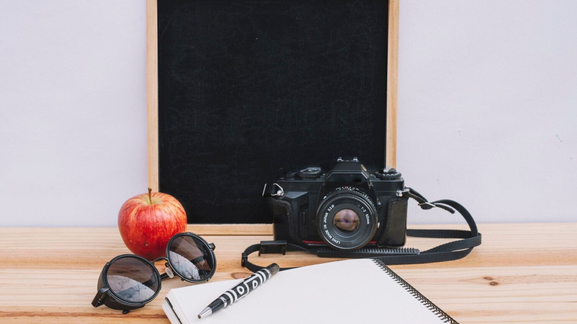 a camera, an apple, a book, a pen and a black board on the wall
